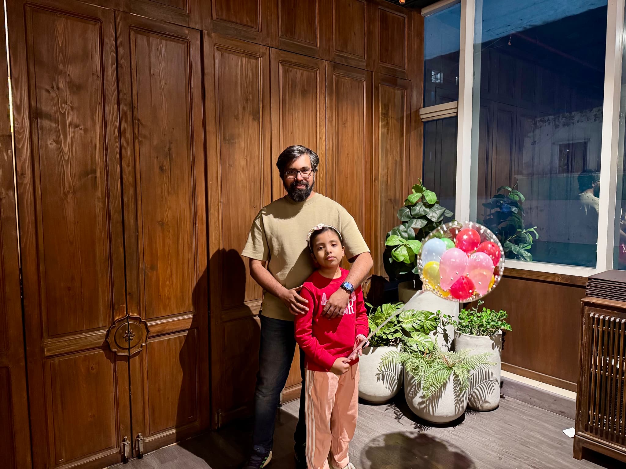 The image features an adult and a child standing in front of what appears to be a wooden structure, possibly inside a building with a high ceiling. The person on the left has dark hair and is wearing a white top, while the person on the right has light-colored hair and is wearing a black shirt. They both seem to be posing for the photo. There are some potted plants visible in the background, as well as what looks like a colorful display of flowers or decorations. The lighting in the image is artificial, with light fixtures hanging from the ceiling providing illumination.