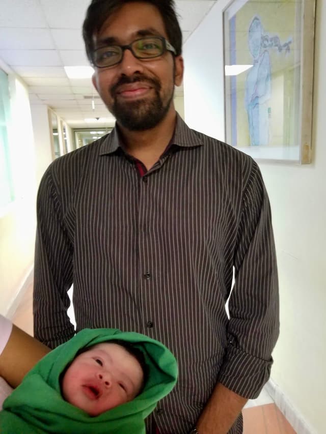 The image depicts a man standing next to a baby swathed in a green blanket, who appears to be at an indoor location. The man is wearing glasses and a patterned shirt. He has dark hair and a beard. Both are looking towards the camera with expressions that could be interpreted as neutral or mildly curious. The background shows a hallway with posters and a painting on the wall.