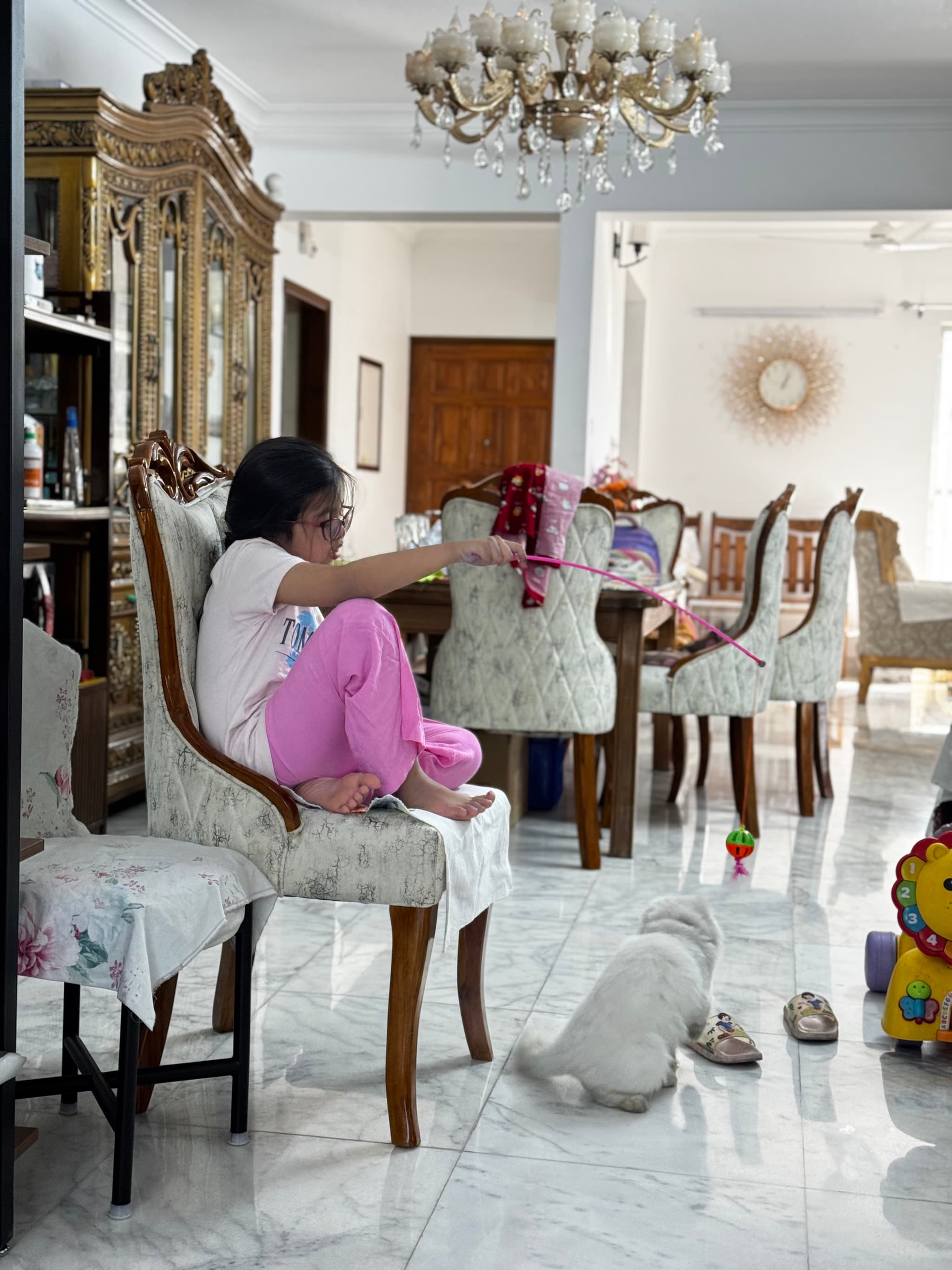 The image depicts an indoor setting with a person sitting on the floor, engrossed in their phone. They are surrounded by several pieces of furniture including chairs and a dining table. In the background, there's a chandelier hanging from the ceiling and what appears to be a decorative object or artwork on the wall. The flooring is tiled, adding to the domestic atmosphere of the scene.
